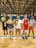 Five young children happily hold out an AFL ball, soccer ball, volleyball, tennis ball and racquet, and small AFL ball.