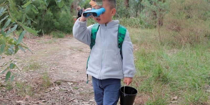 Child walking down path through natural bush setting, holding binoculars up to their eyes