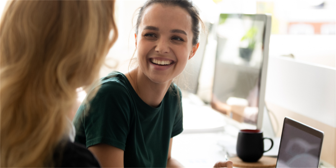 Young person smiling at the person sitting next to them as they both work on the laptop