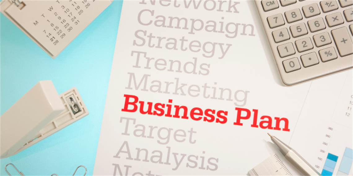 Flat‑lay image of a business planning workspace with a document titled “Business Plan” in red text, surrounded by a calculator, pen, calendar page, paper clips and office stationery on a light blue background