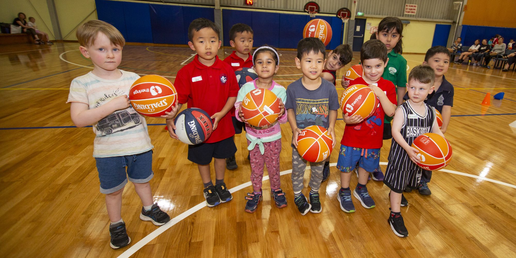 Children holding basketballs during a beginner basketball program on an indoor sports court.