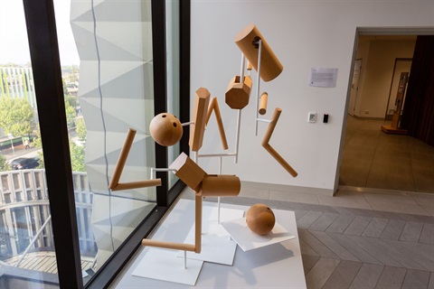 Abstract wooden sculpture composed of geometric shapes and suspended elements displayed on a white plinth beside large windows inside a contemporary building.