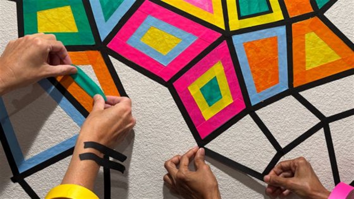 Hands creating colourful geometric artwork using coloured tape.