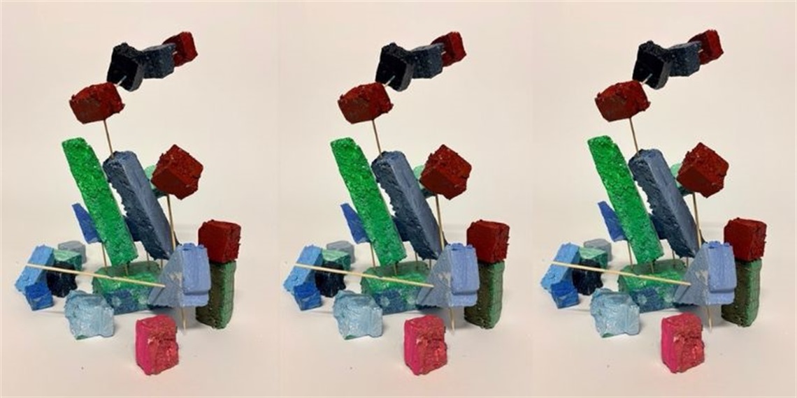 Colourful painted foam blocks and shapes balanced on wooden skewers to create an abstract sculpture against a plain background.