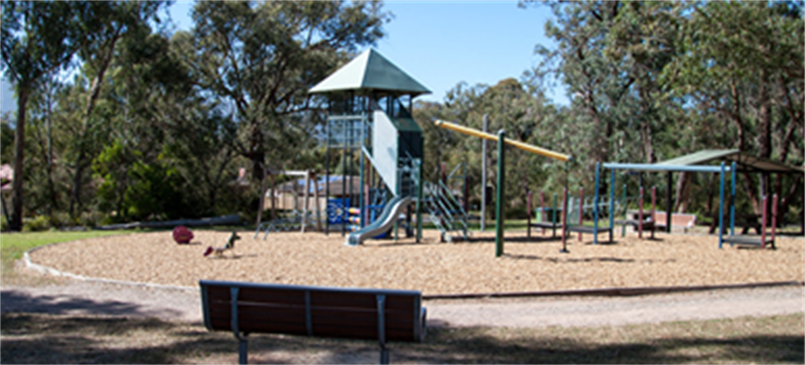 walking path meandering past a colourful playground, with a park bench sitting nearby in the shade