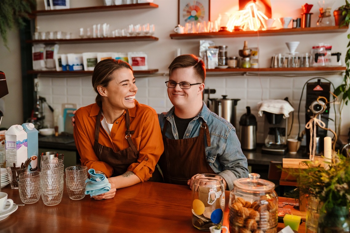 Two people wearing aprons stand behind the counter of a café, surrounded by glasses, coffee-making equipment and shelves stocked with cups and supplies. One person is holding a cloth while both stand side by side in the workspace behind the service counter.