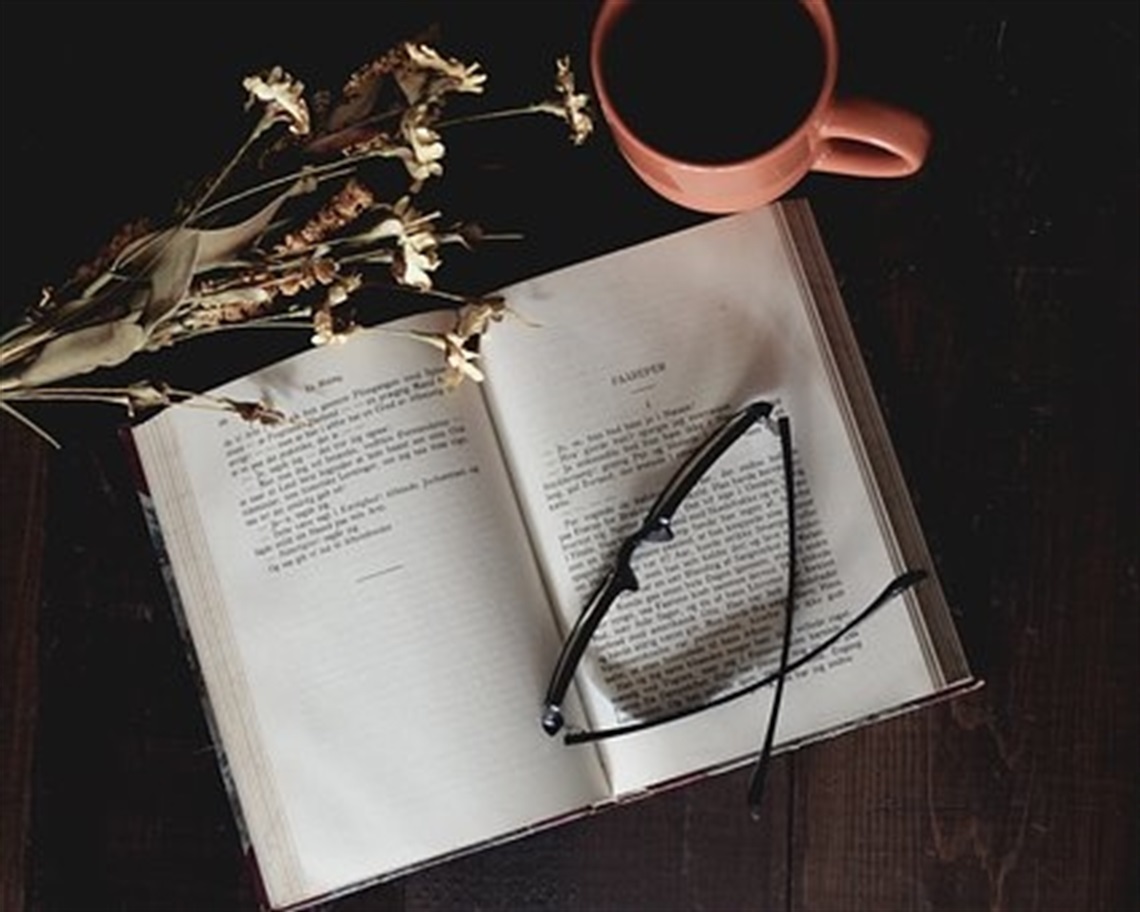book lying open with reading glasses folded on top and dried wildflower lying across the top of the page. A cup of tea or coffee sits nearby.