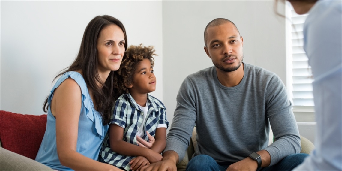 Two parents sitting on a couch talking to someone sitting opposite them. A child sits on their knee and is looking out the window.
