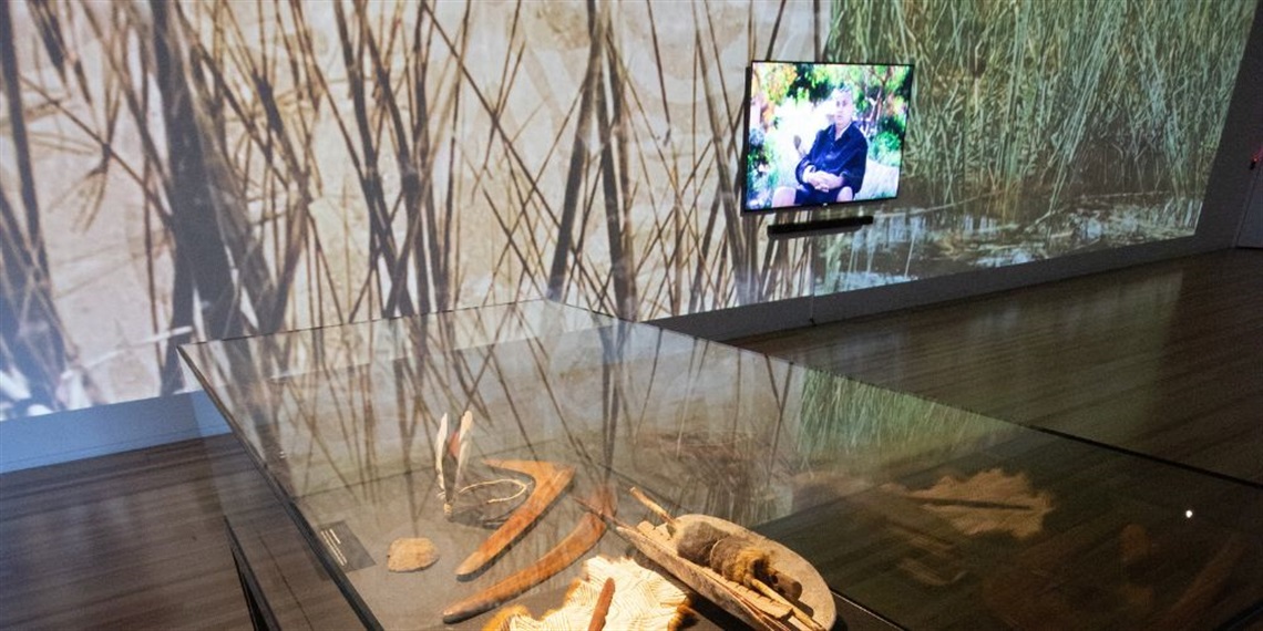 Photo of the 'From our Elders' exhibit showing some First Peoples materials in a glass cabinet while the video plays on the TV in the background
