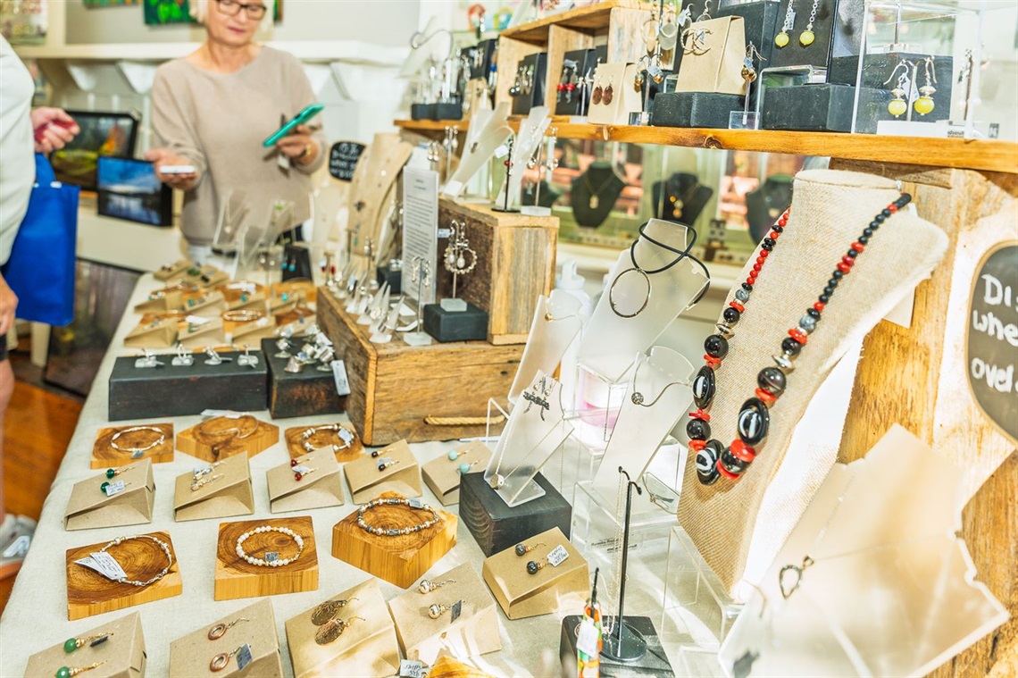 Handmade jewellery displayed on tables and wooden stands at a market stall, including necklaces, earrings, bracelets and rings, while shoppers browse in the background.