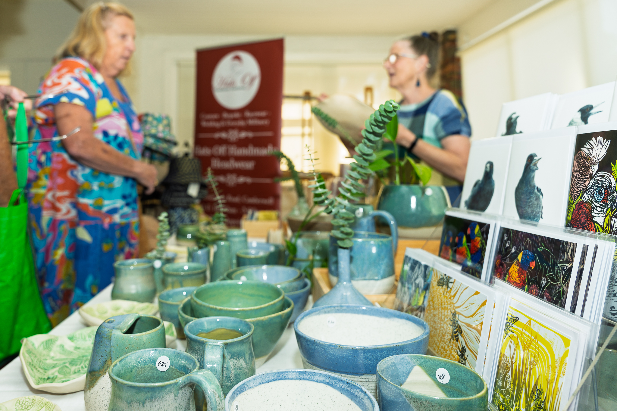 Visitors browse a handmade market stall displaying ceramic bowls, mugs and vases alongside art prints and greeting cards, with stallholders chatting in the background.