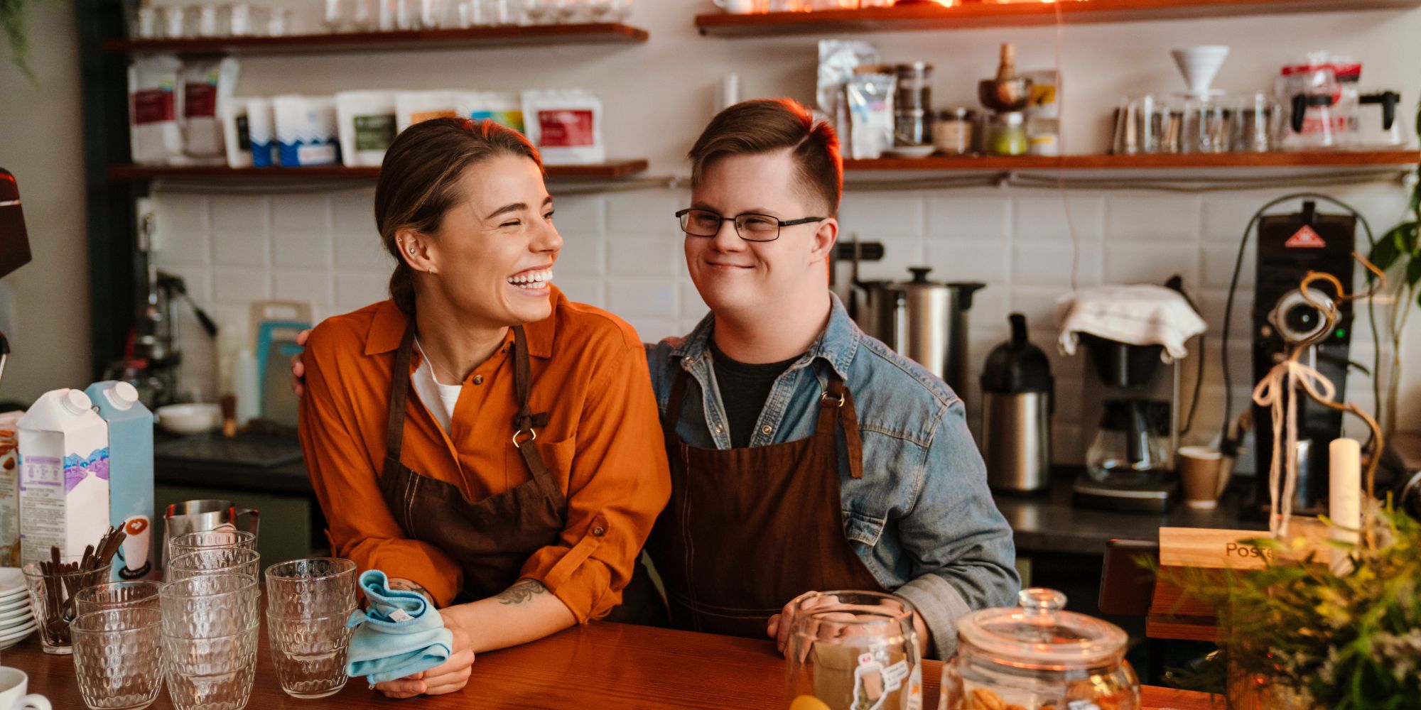 Two people wearing aprons stand behind a café counter surrounded by glasses, coffee-making equipment and food preparation items.