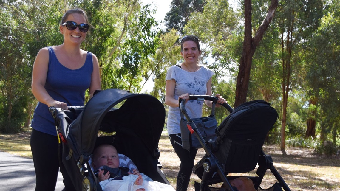 Two mothers walking along a nature trail, pushing their babies in prams