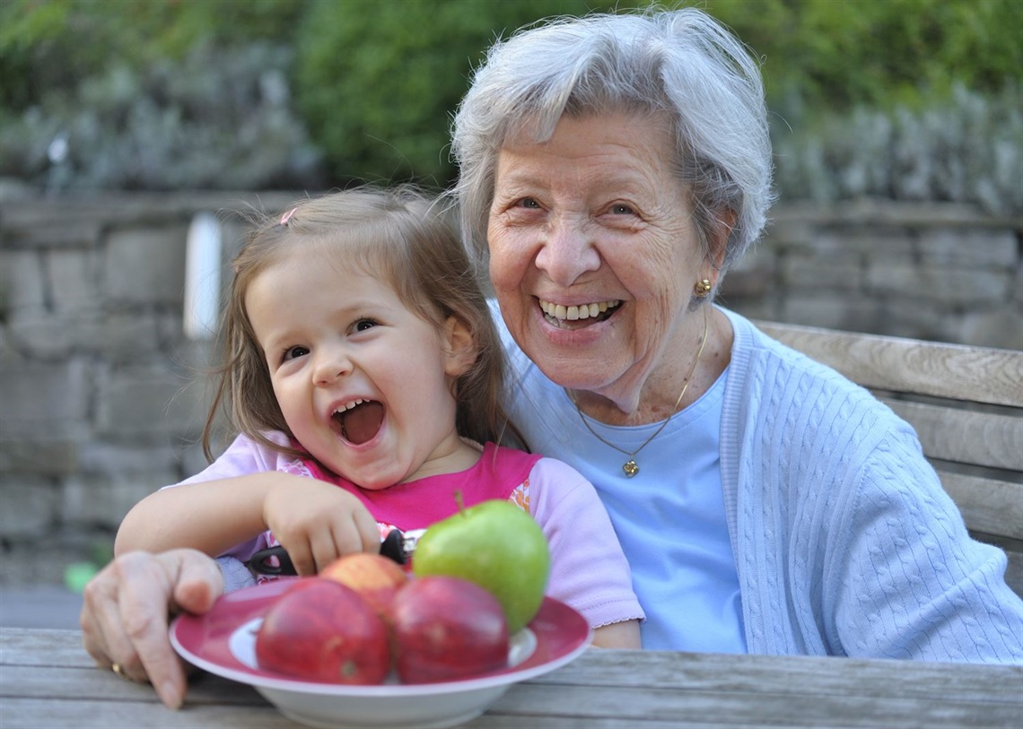 An older adult and a young child sitting together on a wooden bench outdoors, holding a plate with apples and a pear. A stone wall and greenery are in the background.