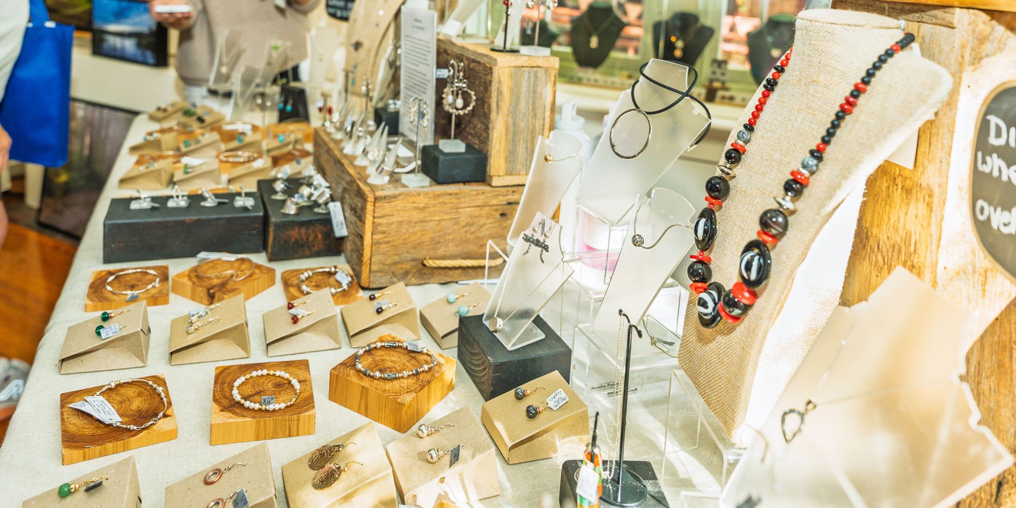 Handmade jewellery displayed on tables and wooden stands at a market stall, including necklaces, earrings, bracelets and rings, while shoppers browse in the background.