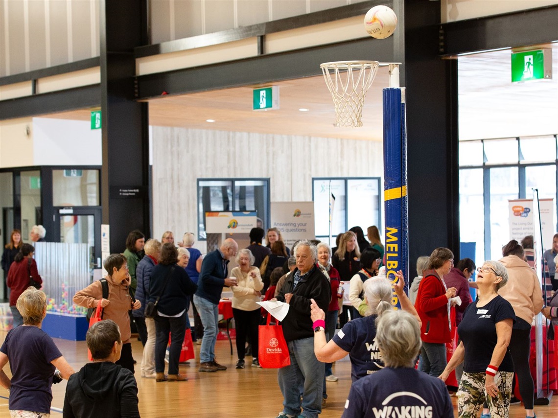 Adults participating in a walking netball activity at the Movement and Positive Ageing Expo 2025, with attendees and information stalls in the background.