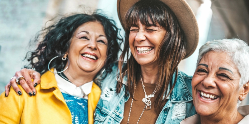 Three people standing arm in arm, wearing colourful casual clothing, in a close-up outdoor photo.