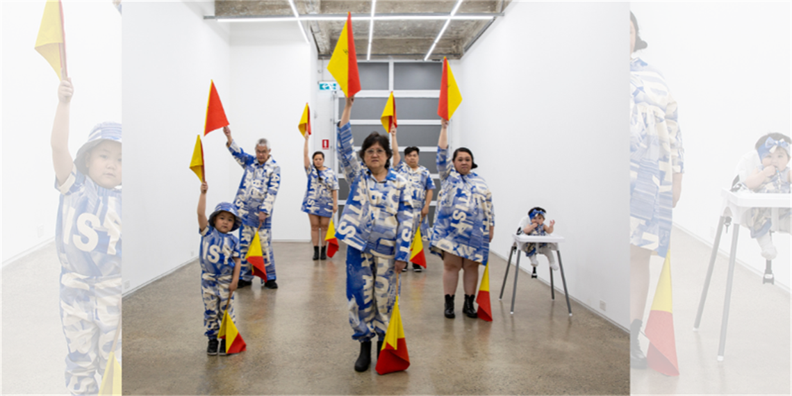 Group of adults and children in a gallery space holding red and yellow semaphore flags while wearing matching blue and white costumes.