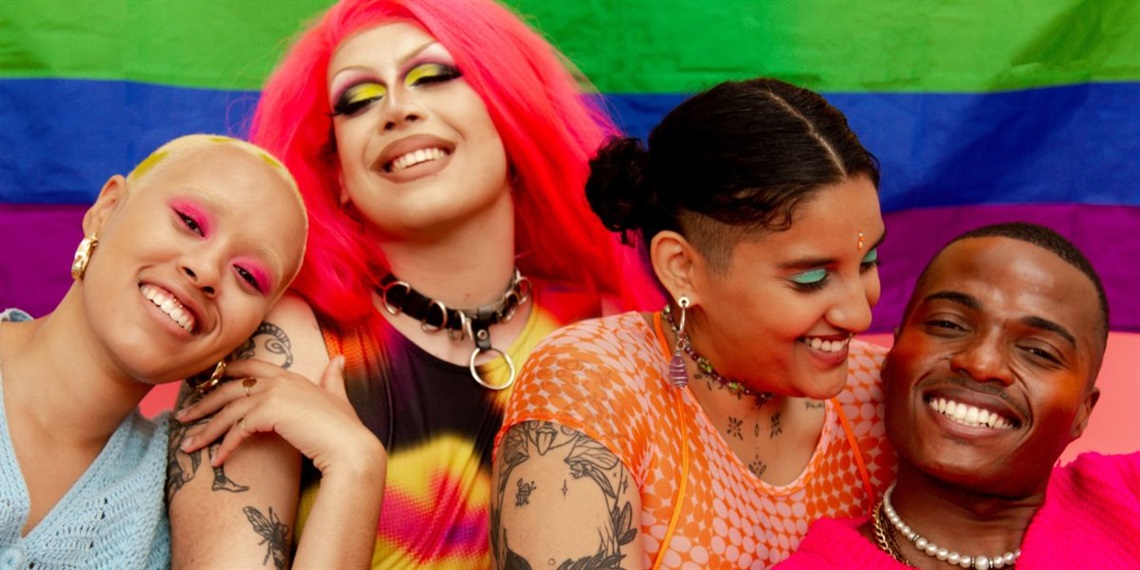 Four people in bright clothes and makeup sitting in front of a Pride flag.