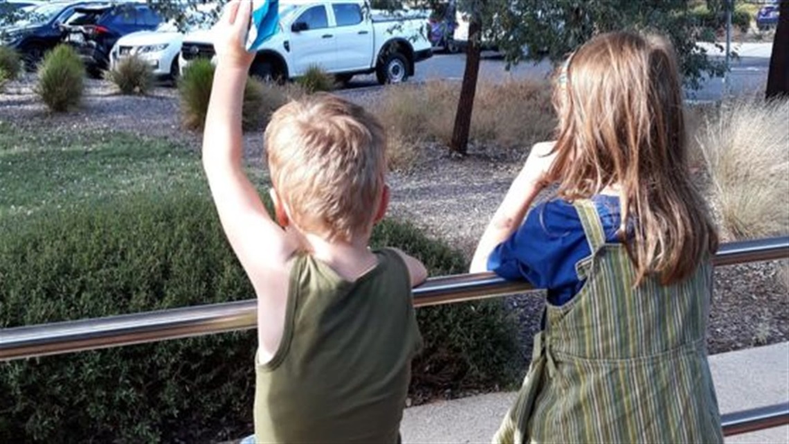 Two children sit on outdoor railings overlooking a garden area.