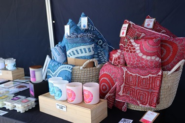 Market stall displaying patterned cushions, woven baskets and scented candles on a table.