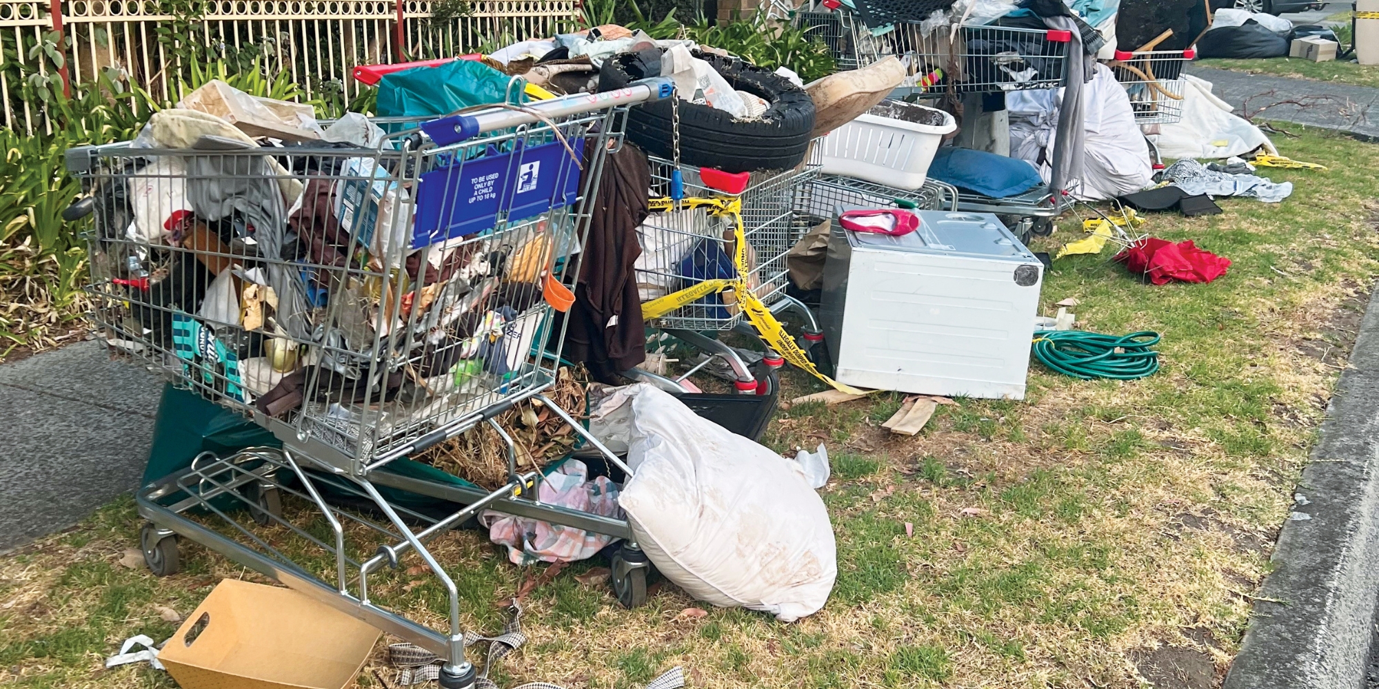 Several shopping trolleys filled with dumped household rubbish on a grass verge beside a footpath.